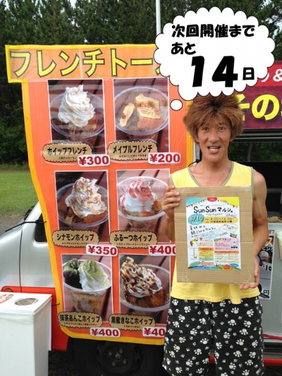 キッチンカーのメニュー看板の前で、マルシェの告知プラカードを持つ初代店主